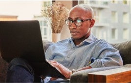 A recruiter is sitting on a couch and working on a laptop. He has dark glasses and is wearing a blue shirt.